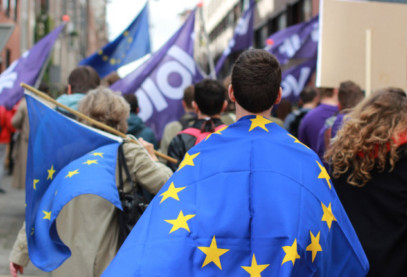 A man in the foreground wearing a European Flag. More European flags are seen in the background with people also waving Volt flags.