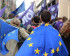 A man in the foreground wearing a European Flag. More European flags are seen in the background with people also waving Volt flags.