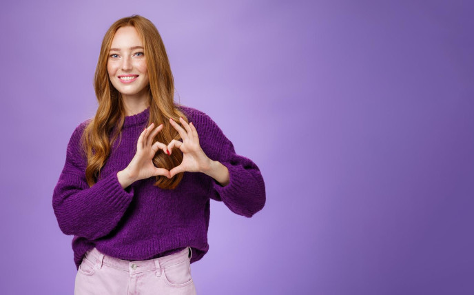 Woman with ginger hair and a purple jumper is forming a heart with her fingers