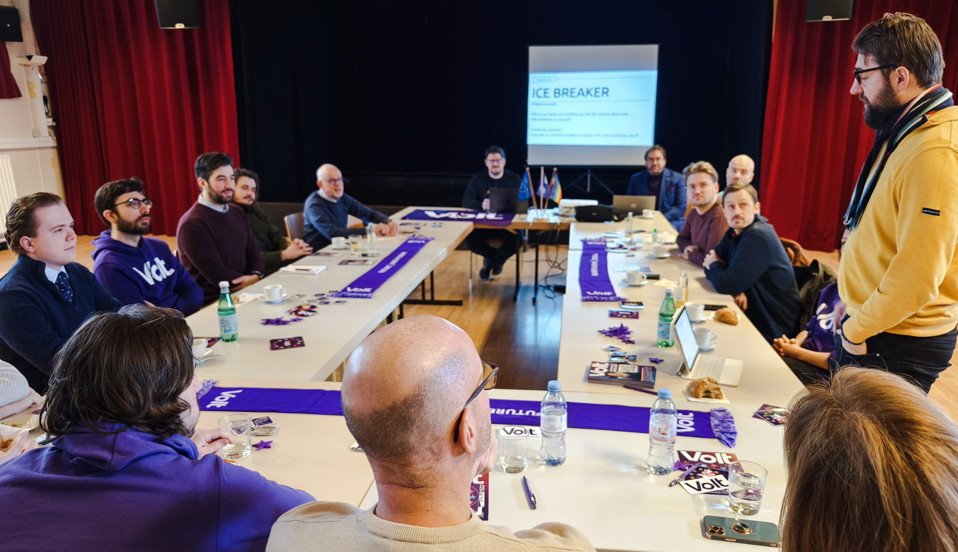 Tables arranged in a square, Volters are sitting at the table, in the background you can see a stage with a screen on which a presentation is running. On the right, George Penn is standing and saying something.